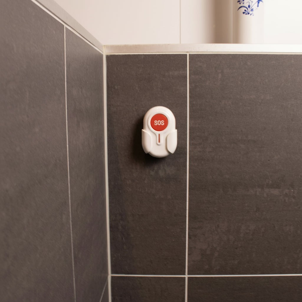 DigiWelzijn Aupero ovale waterbestendige SOS-alarmknop gemonteerd aan een grijze badkamertegelmuur
